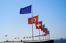 Foto av piren i Helsingborgs hamn. Man ser flera olika flaggor som EU-flaggan, Turkiets flagga, Schweiz flagga med flera mot en klarblå himmel. I bakgrunden skymtar Danmarks kustremsa.